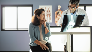 Middle-aged Hispanic woman in a medical office examining a knee X-ray showing signs of osteoarthritis, including joint space narrowing and osteophytes. A Hispanic physician points to the image, illustrating the diagnostic process for osteoarthritis and joint degeneration.